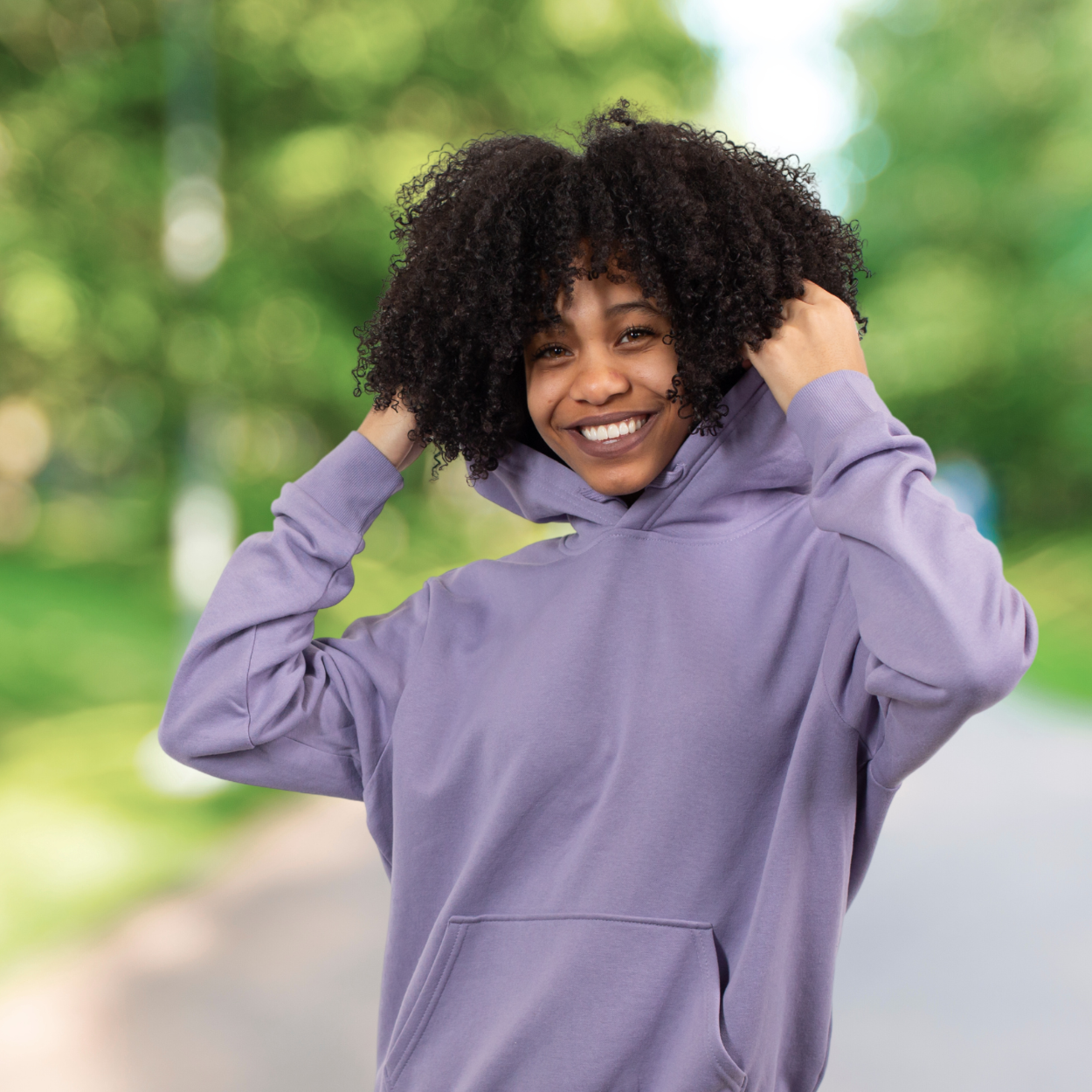 MR Spravato Purple jogger Smiling person in a light purple hoodie adjusting the hood while standing on a sunlit path surrounded by greenery. Featured on the Mind Rejuvenation Home page to reflect confidence, joy, and the uplifting power of nature in mental wellness.