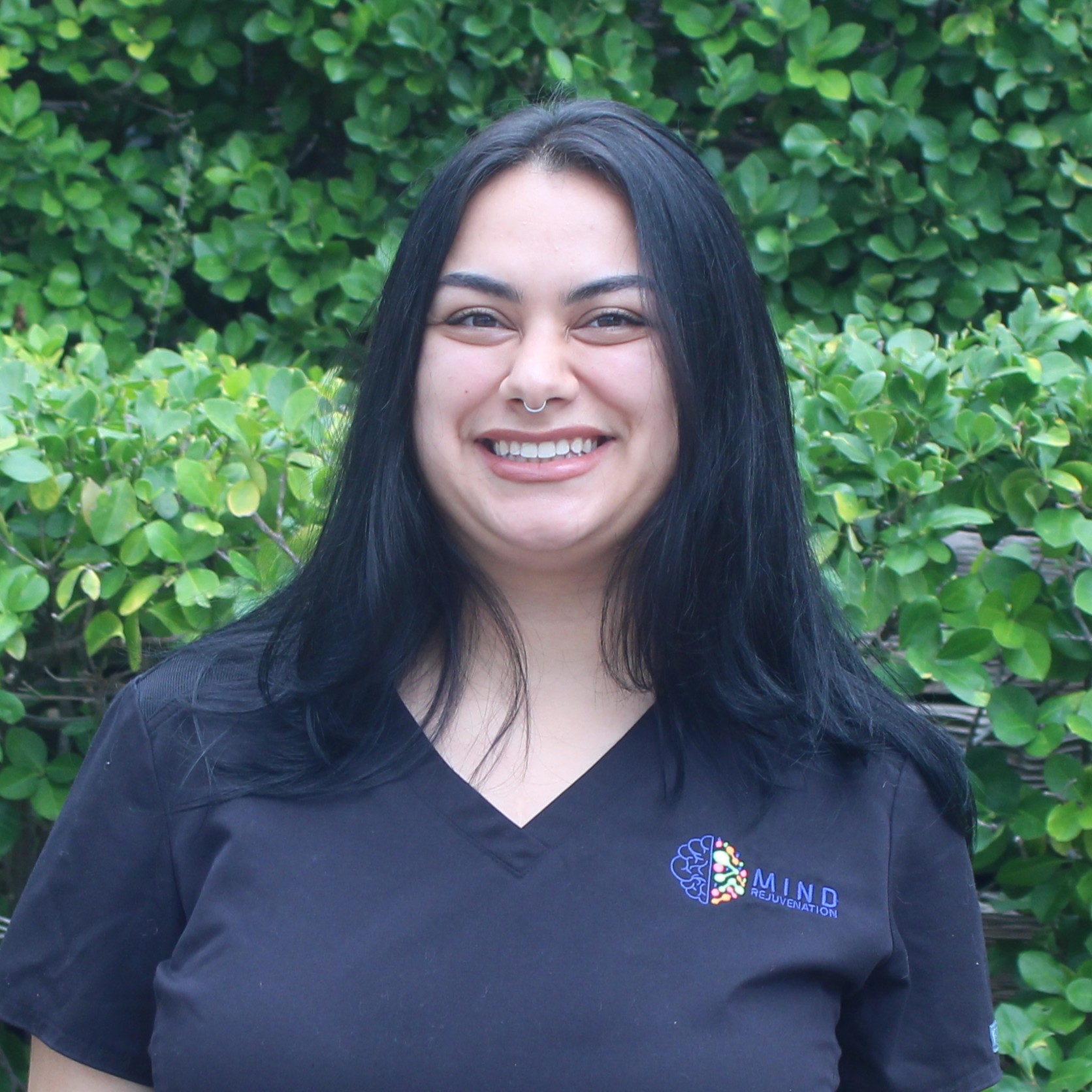 Breanna Page, Patient Coordinator at Mind Rejuvenation, smiling outdoors in branded attire with a green hedge backdrop. Featured on the Meet Our Team page to reflect warmth, organization, and a welcoming presence in patient support.