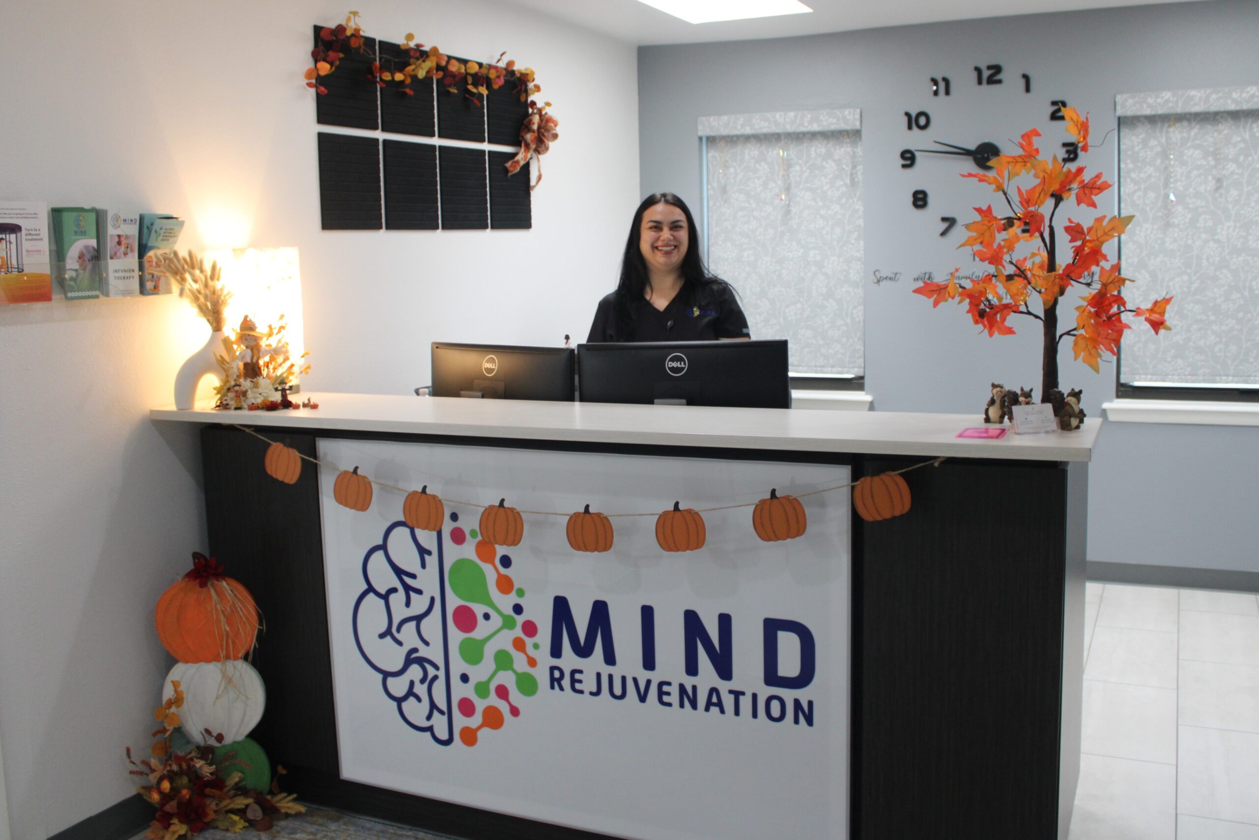 Front Desk Smiling receptionist behind the Mind Rejuvenation front desk, surrounded by autumn-themed decor and wellness branding. Featured on the Home page to reflect warmth, professionalism, and a welcoming check-in experience for mental health care.