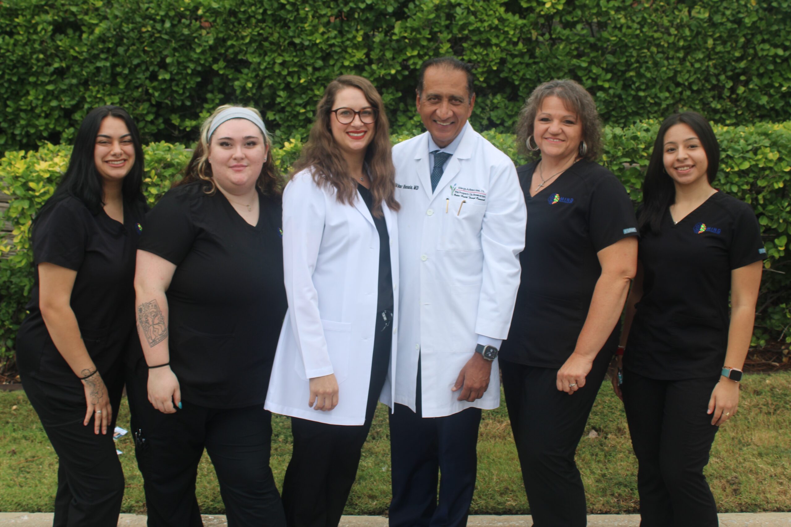MR Team1 Group of six smiling healthcare professionals—four in black scrubs and two in white lab coats—standing outdoors in front of a green hedge. Featured on the Mind Rejuvenation Home page to highlight compassionate care, clinical expertise, and a welcoming wellness team.