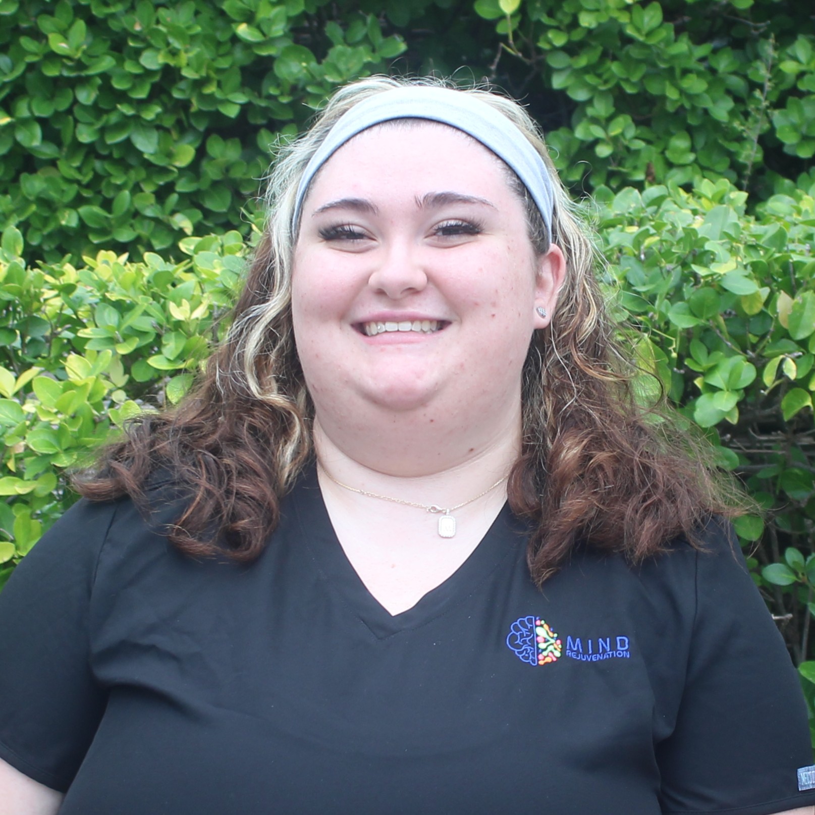 Rosy, Certified Nursing Assistant (CNA) at Mind Rejuvenation, smiling in branded attire outdoors with a green hedge backdrop. Featured on the Meet Our Team page to reflect gentle care, clinical support, and a warm, welcoming presence in patient wellness.