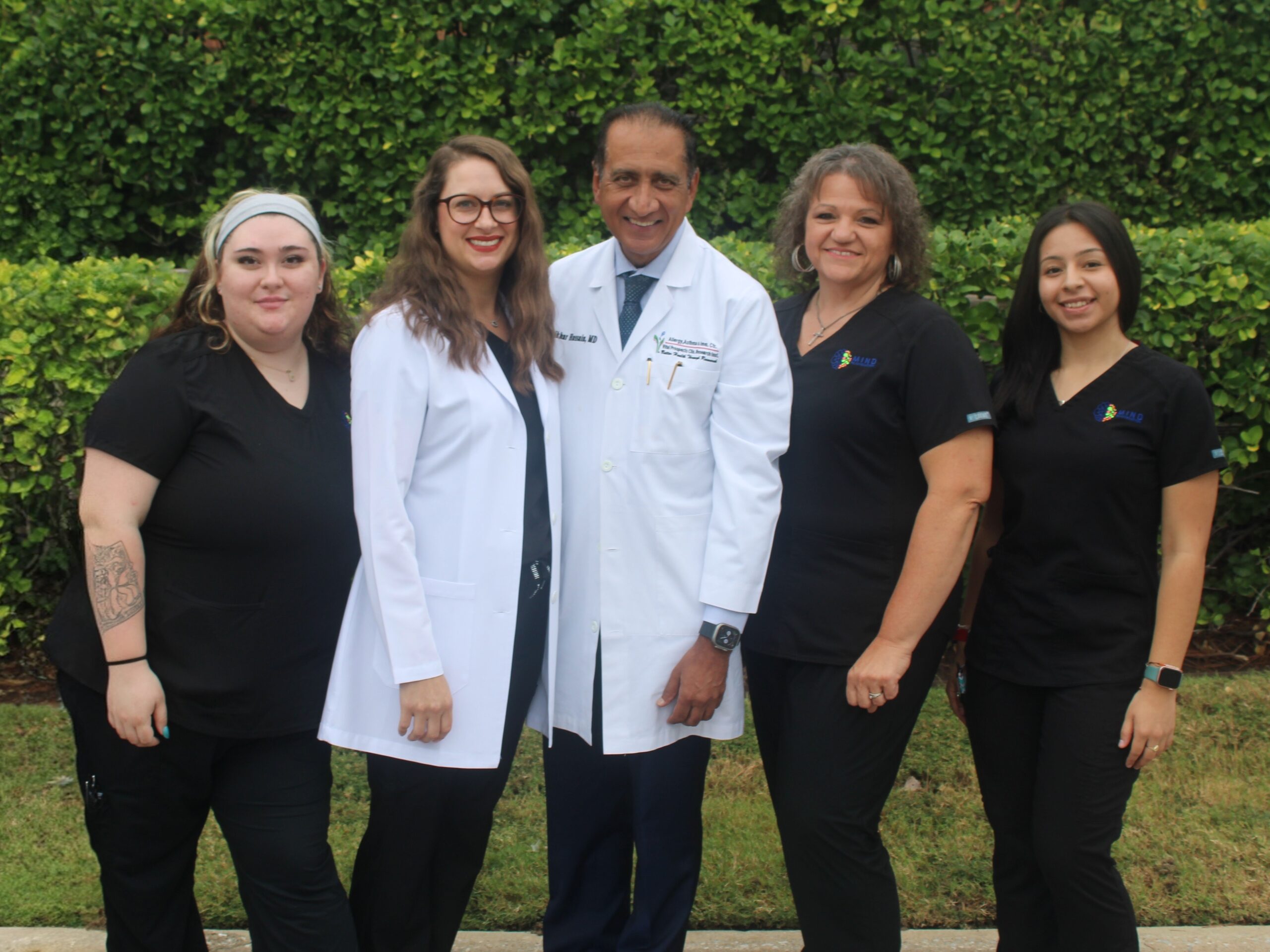 Group of five smiling healthcare professionals—four in black scrubs and two in white lab coats—standing outdoors in front of a green hedge. Featured on the Mind Rejuvenation Home page to highlight compassionate care, clinical expertise, and a welcoming wellness team.